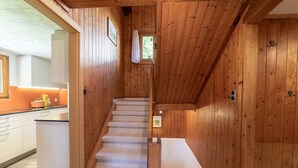 Un interior de madera con una escalera que conduce a un segundo piso, una zona de cocina con armarios blancos y un ventanal con vistas a la vegetación.