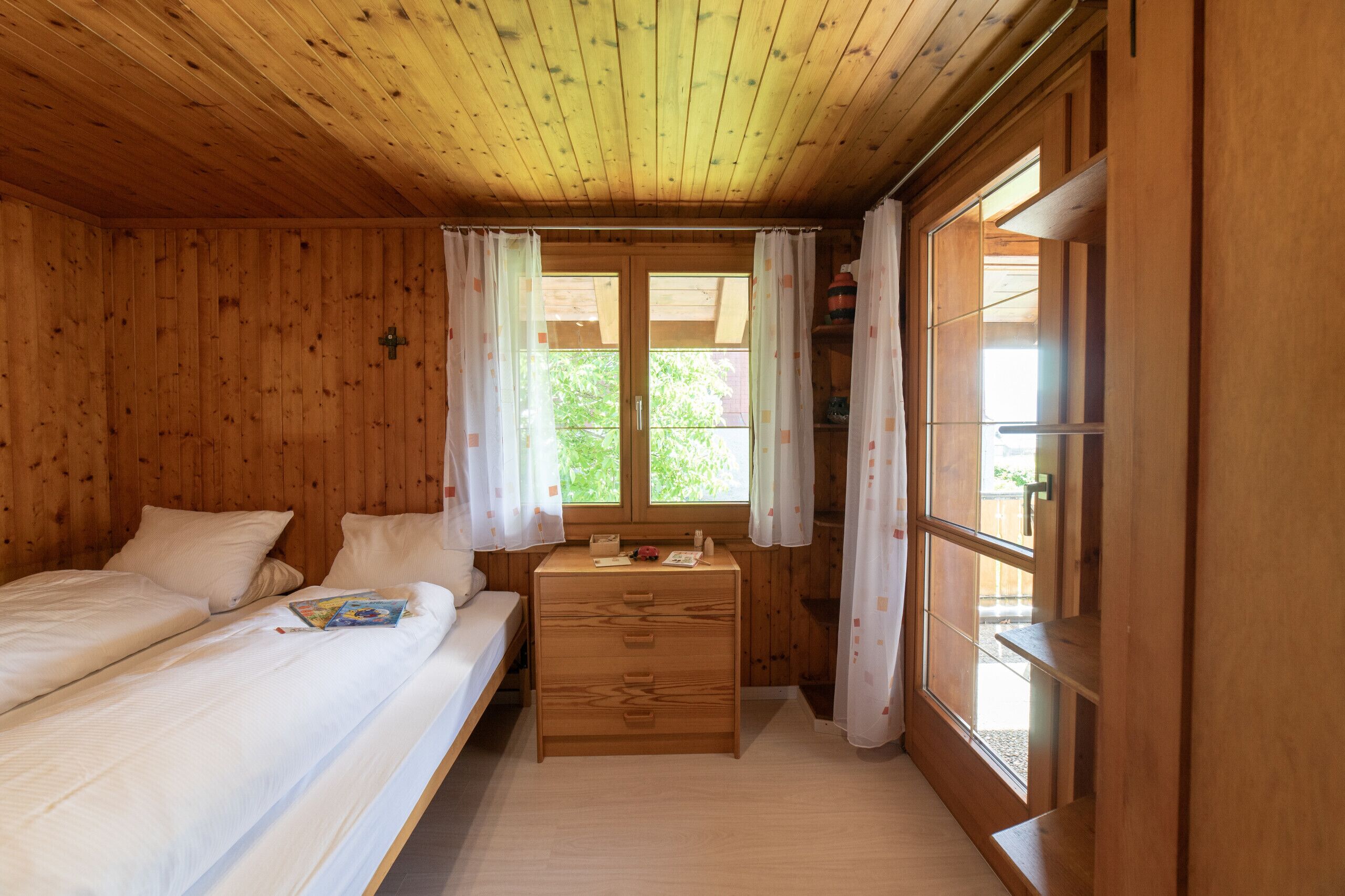 A wooden room with two beds, a small wooden dresser, and a window with curtains.