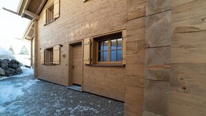 Un edificio de madera con entrada empedrada de piedra, contraventanas de madera en las ventanas y una pared de piedra al fondo.