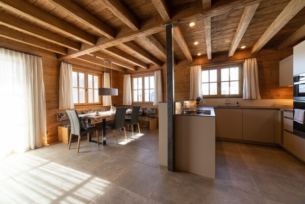 Eine geräumige Küche mit Holzbalken, ein Essbereich mit Tisch und Stühlen sowie große Fenster mit Vorhängen.