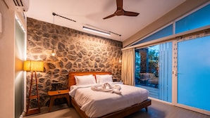 A bedroom with a stone wall, a wooden bed, a bedside table, a lamp, and a ceiling fan. - Basalt Paradise Resort And Spa (Halol)