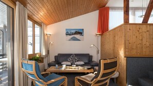 Um quarto aconchegante com teto de madeira, um sofá, duas cadeiras de madeira, uma mesa de centro e uma janela com cortinas.