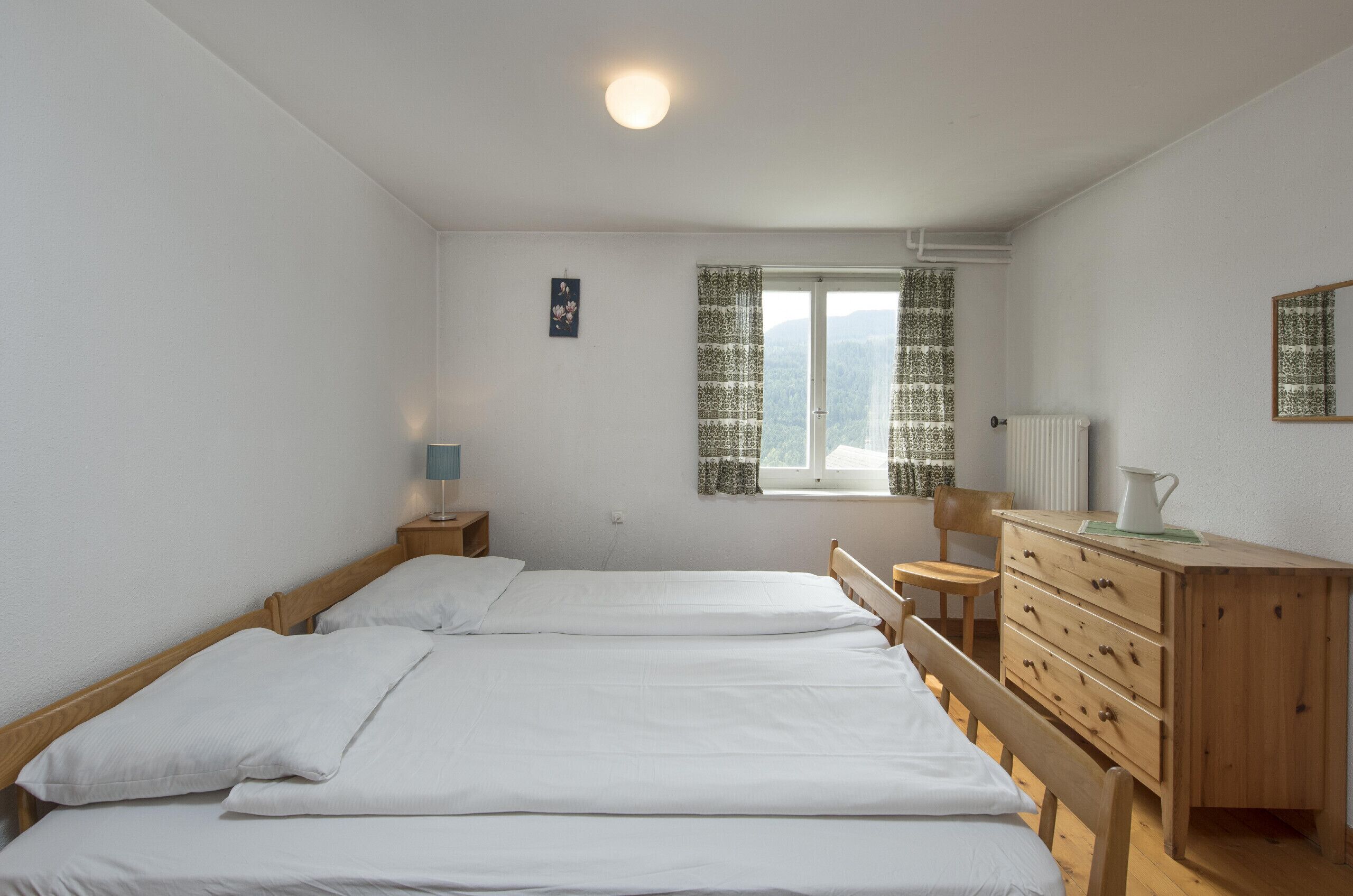 Une chambre avec deux lits simples, une commode en bois, une chaise, un radiateur et une fenêtre avec des rideaux.