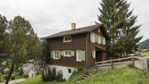 Une maison à deux étages, avec un toit en pente, des volets verts et une clôture en bois.