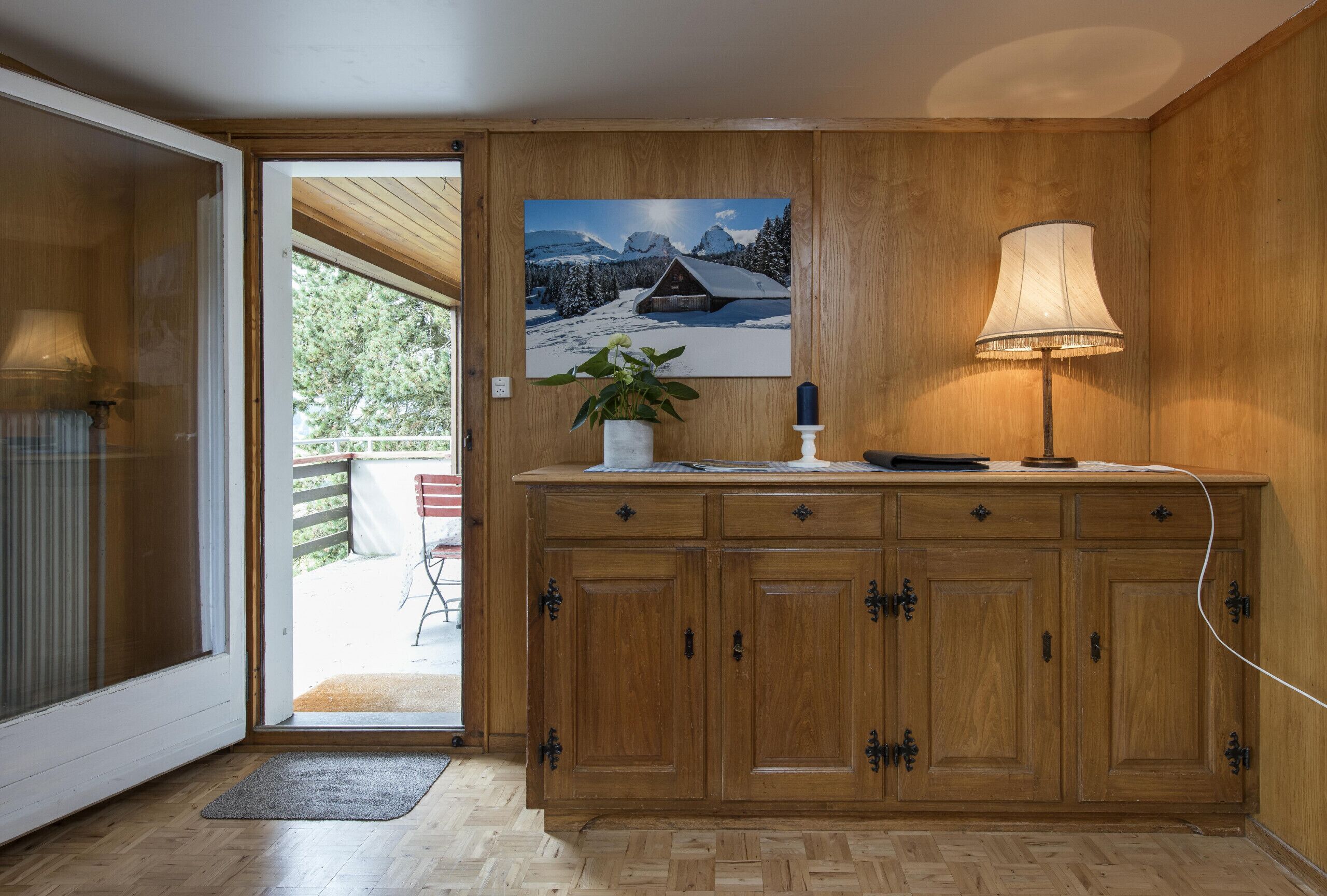 Ein Holzschrank mit einem Gemälde einer verschneiten Berglandschaft, einer Lampe und einer Topfpflanze.