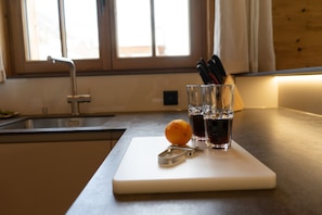 Una encimera de cocina con tabla de cortar, un vaso de agua, un vaso de líquido oscuro y una naranja.