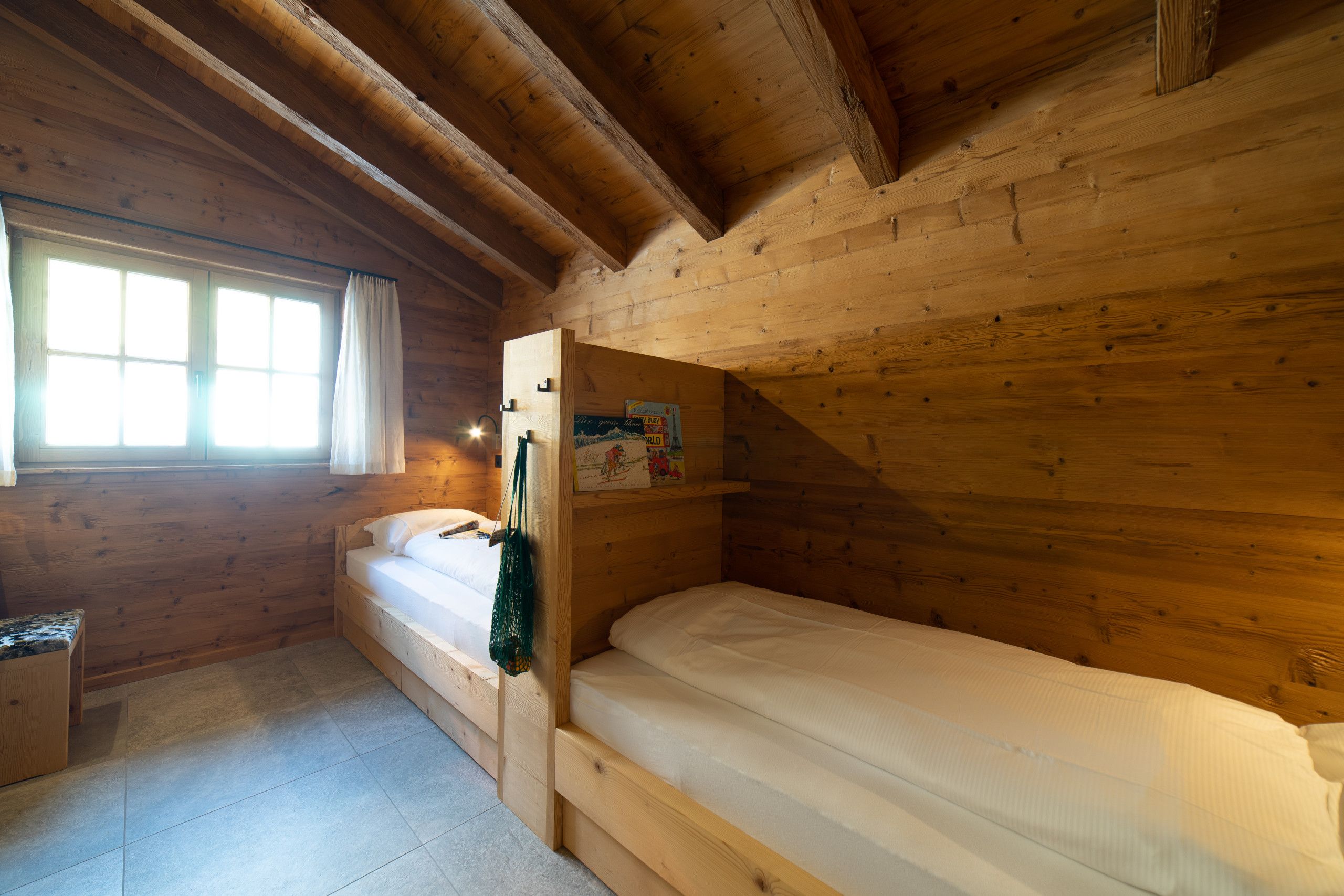A small, wooden cabin room with a single bed, a window, and a shelf with items.