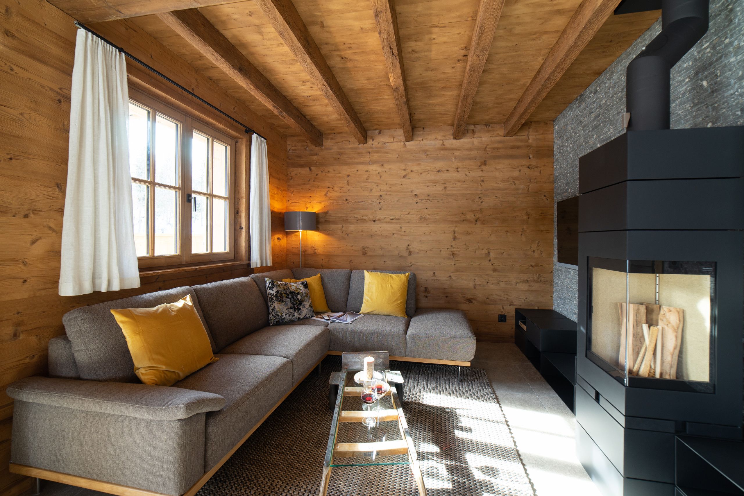 A cozy log cabin interior with a sectional sofa, a glass coffee table, and a wood-burning stove.