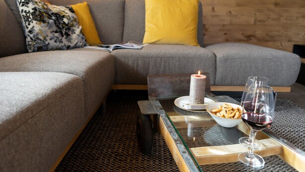 En moderne stue med en grå sofa, gule puder, et glasbord, et tændt stearinlys, en skål med snacks og et glas rødvin.