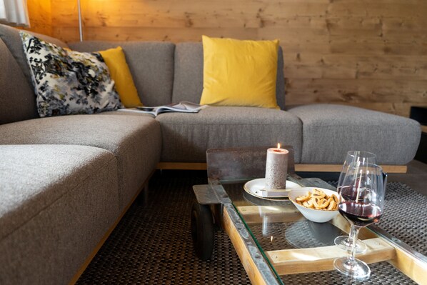 Una sala de estar moderna con un sofá gris, cojines amarillos, una mesa de centro de vidrio, una vela encendida, un bol de aperitivos y una copa de vino tinto.