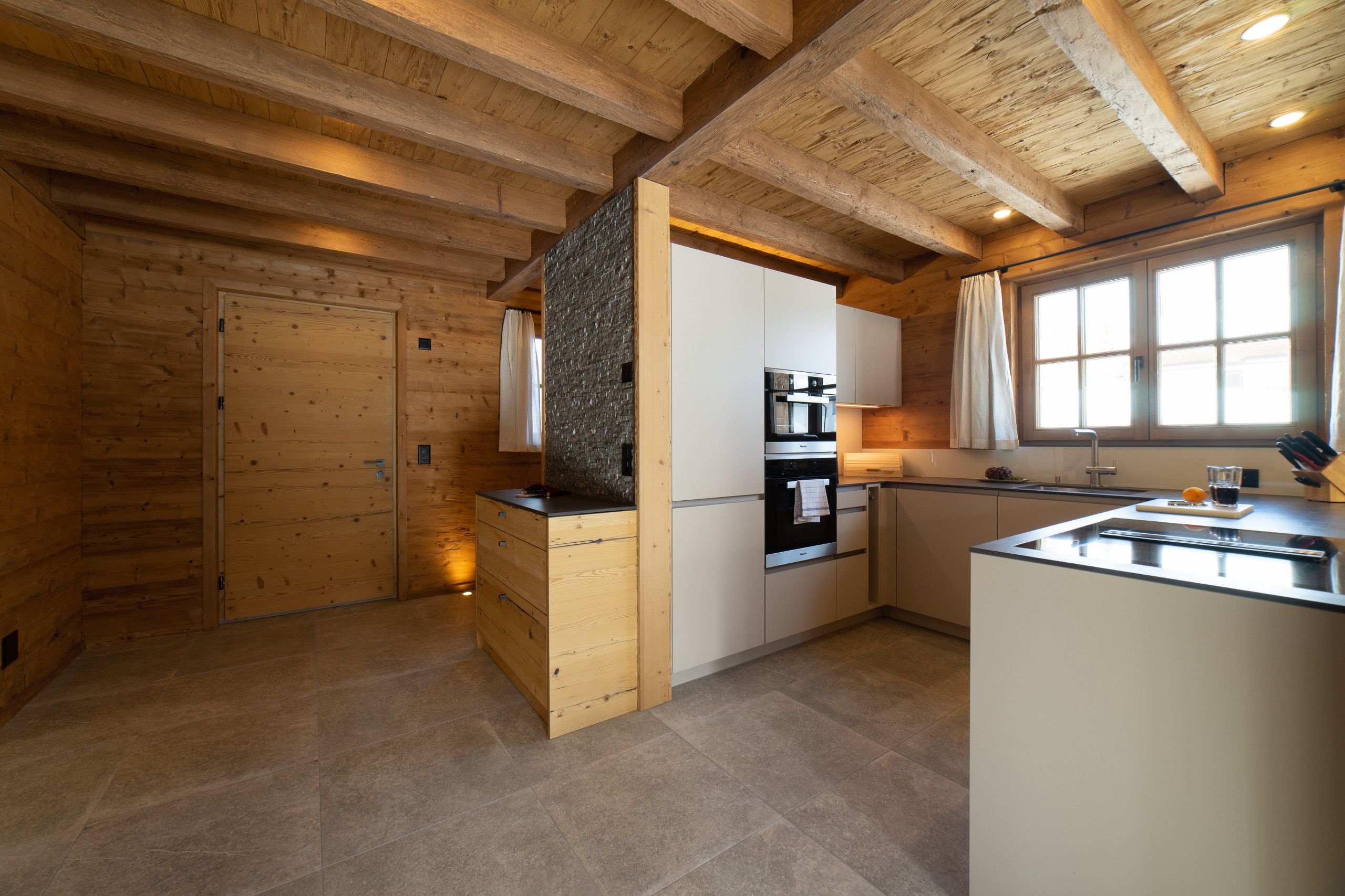 A modern kitchen with wooden beams, a stone accent wall, and a large window.