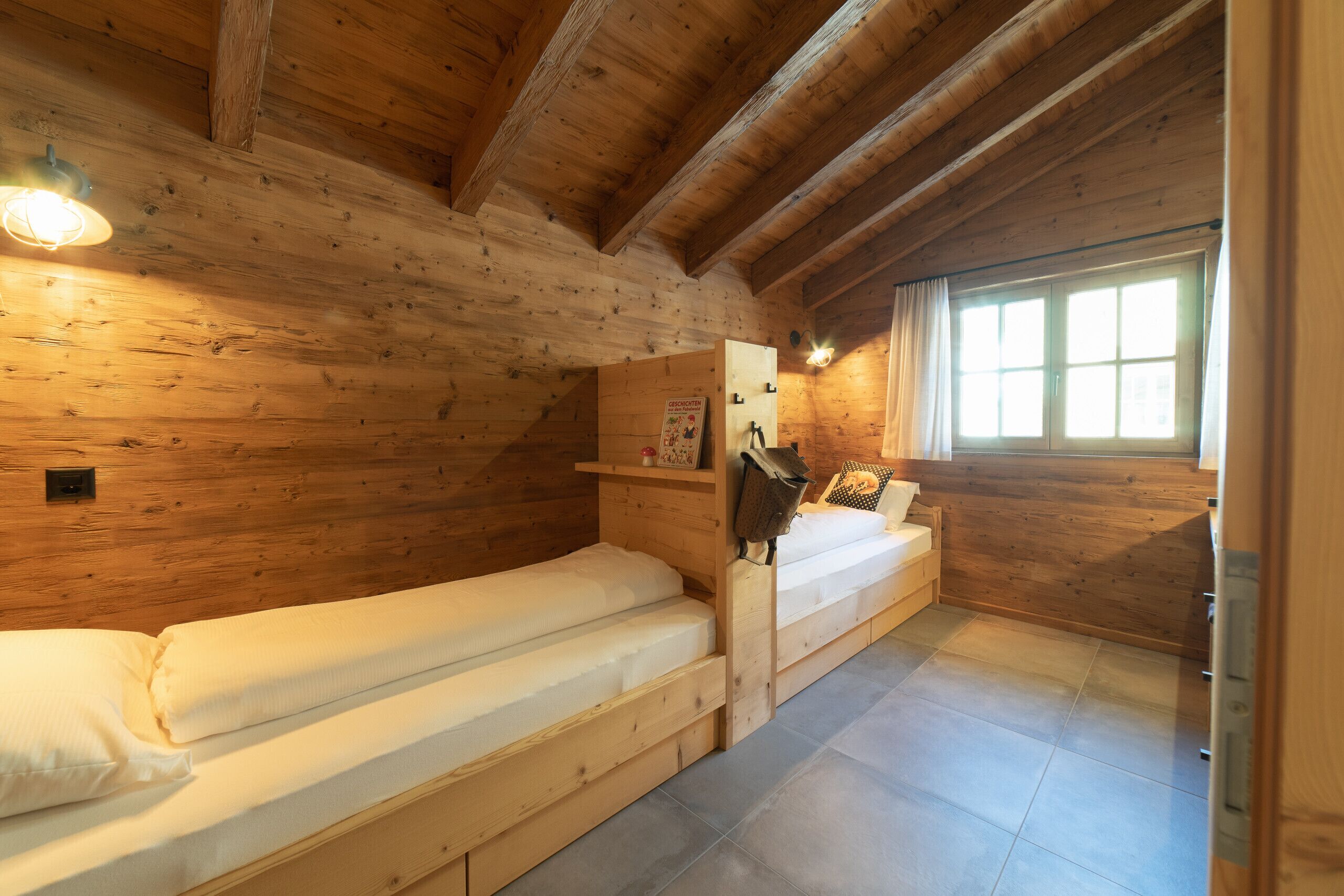 Ein Zimmer einer Holzhütte mit zwei Betten, einem Fenster und einem Regal mit Gegenständen.