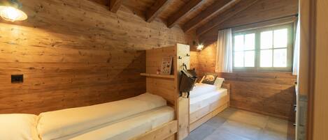 Ein Zimmer einer Holzhütte mit zwei Betten, einem Fenster und einem Regal mit Gegenständen.