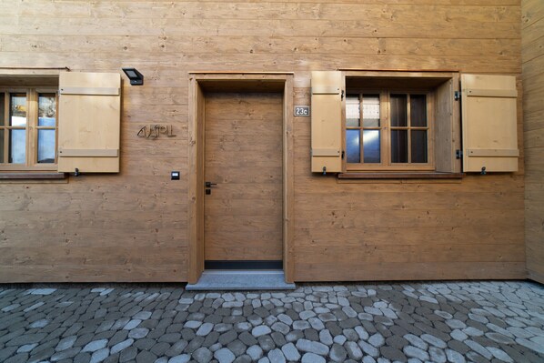 Un edificio de madera con una puerta central, dos ventanas con persianas y un suelo de adoquines.
