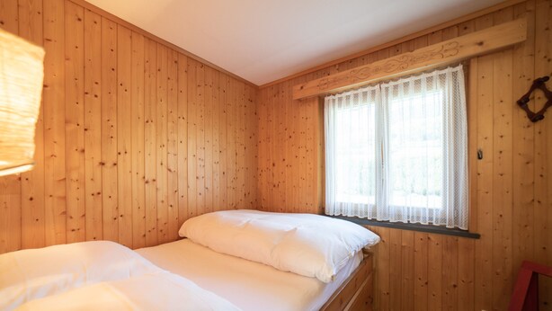 Uma cama de solteiro num quarto com paredes de madeira e uma janela com cortinas de renda.