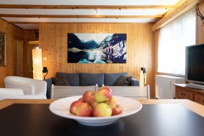 Una sala moderna con detalles de madera, un cuadro grande de un paisaje montañoso, un sofá, un televisor y una mesa con manzanas.