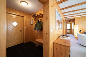 A wooden interior with a door, a bench, a coat rack, and a small table with a fruit basket.