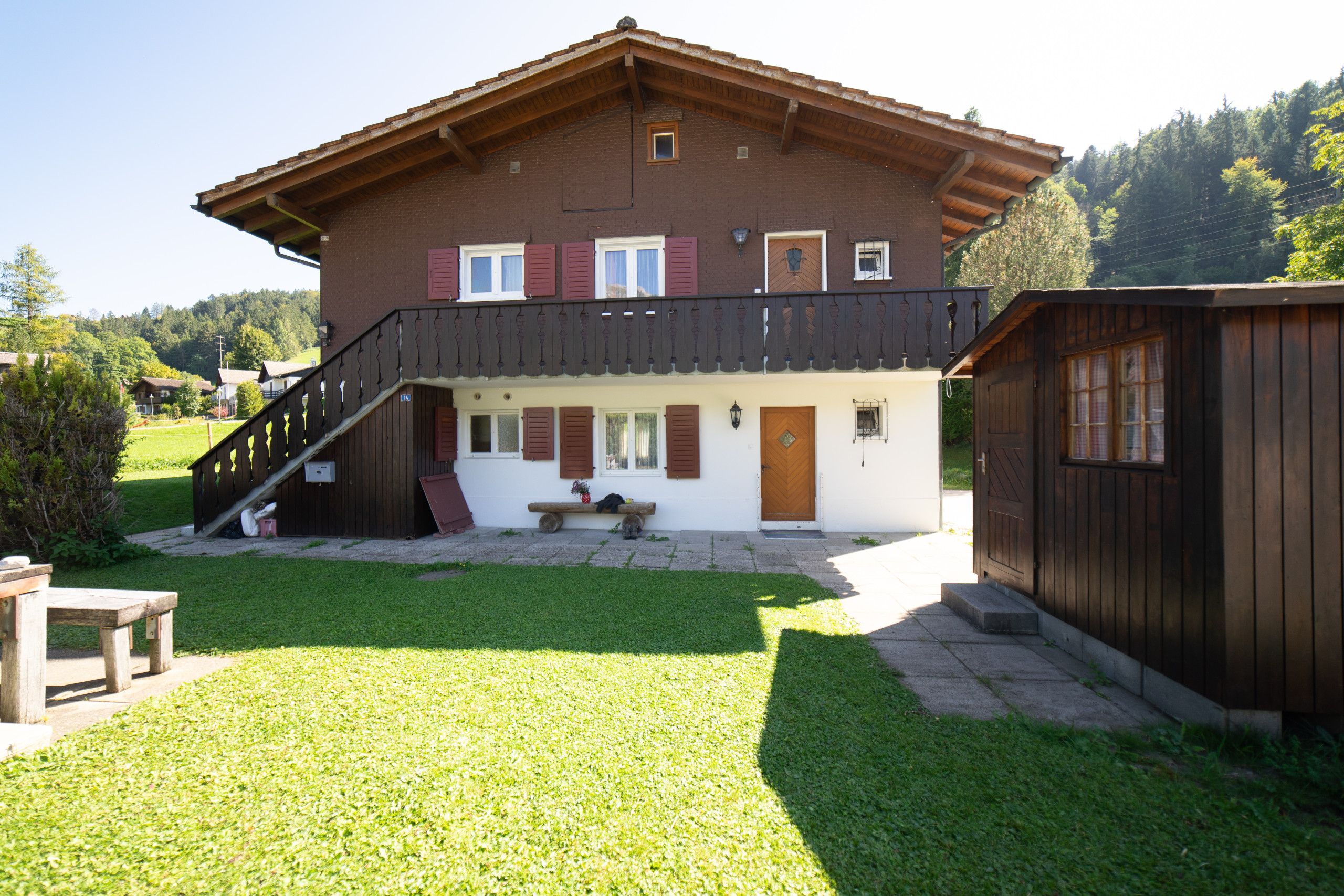Un chalet de dos pisos con un balcón, una terraza de madera y un cobertizo pequeño.