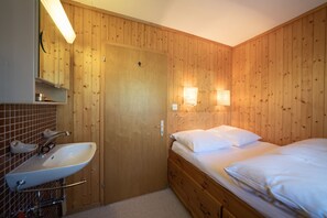 Una habitación con paredes de paneles de madera, un lavabo blanco, un espejo, una cama con ropa de cama blanca y dos lámparas fijadas a la pared.