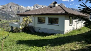 Una pequeña casa de un solo piso con techo de tejas, ubicada en una pendiente cubierta de hierba con montañas al fondo.