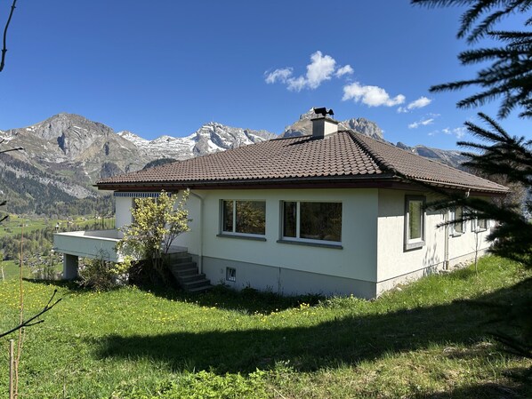 Una pequeña casa de un solo piso con techo de tejas, ubicada en una pendiente cubierta de hierba con montañas al fondo.