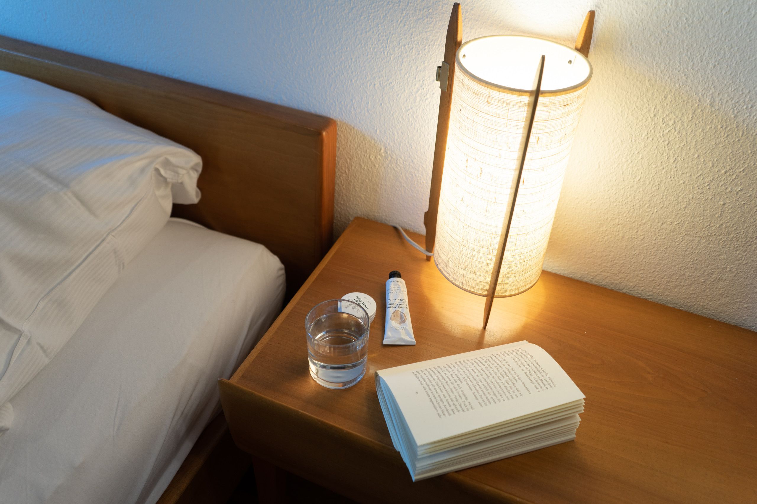 A neatly made bed with white linens, a bedside table with a lamp, a glass of water, a tube of cream, and a stack of books.