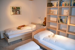 Un dormitorio con dos camas individuales, una mesita de noche con lámpara, una estantería y un cuadro enmarcado en la pared.