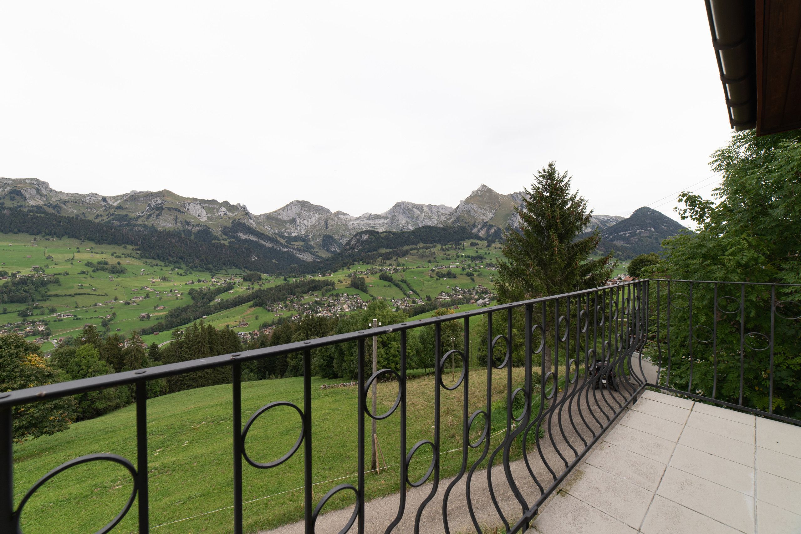 Un balcon doté d’une balustrade en métal, d’où l’on peut admirer un paysage montagneux parsemé de maisons.
