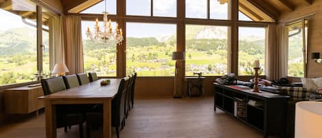 Una habitación amplia con techo de madera alto, ventanas grandes que ofrecen vistas a un paisaje montañoso, una mesa de comedor con sillas y una zona de estar acogedora con un sofá y una mesita.