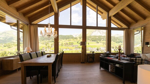 A spacious room with a large dining table, a cozy seating area, and a view of a mountainous landscape through large windows. - Primula 4.5 room chalet next to the slopes (Wildhaus)