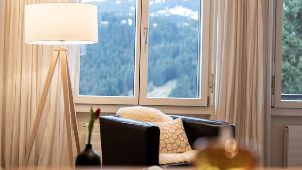 A cozy living room with a sofa, a lamp, and a view of a forest through large windows.