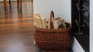 Una esquina de una habitación con una cesta de mimbre llena de leña sobre un banco oscuro, una mesa de comedor de madera con sillas, y una pared negra con una ventana y una tubería negra.