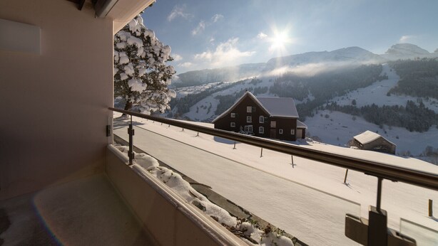 A balcony with a view of a snow-covered landscape, including a house and mountains.