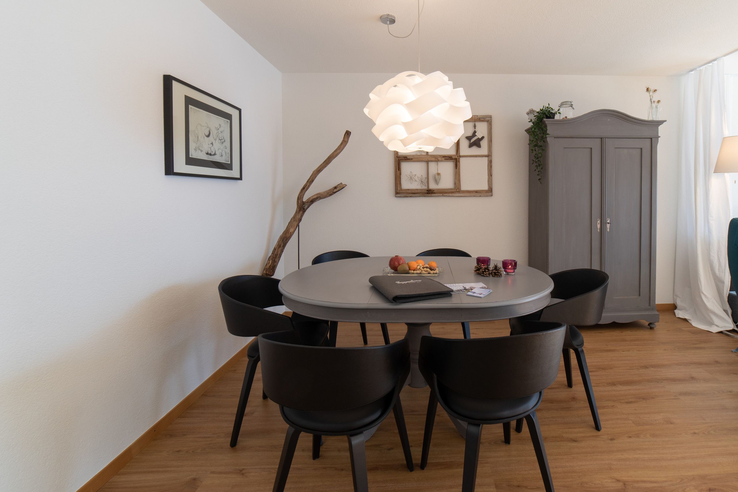 Un comedor con una mesa redonda, sillas negras, una lámpara colgante, un cuadro enmarcado, una rama decorativa y un armario.