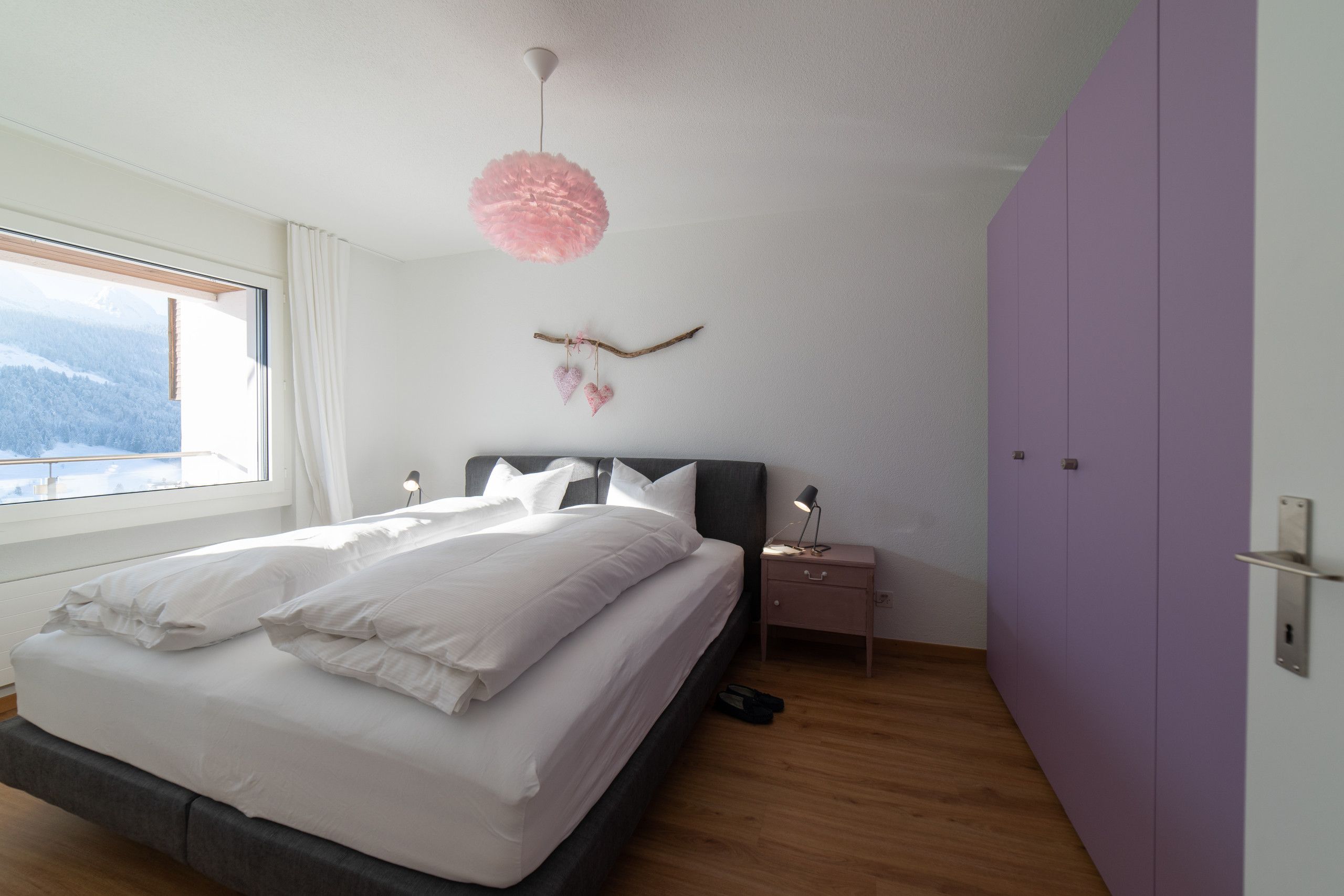 Een slaapkamer met een bed, nachtkastje, lamp, raam en uitzicht op bergen.
