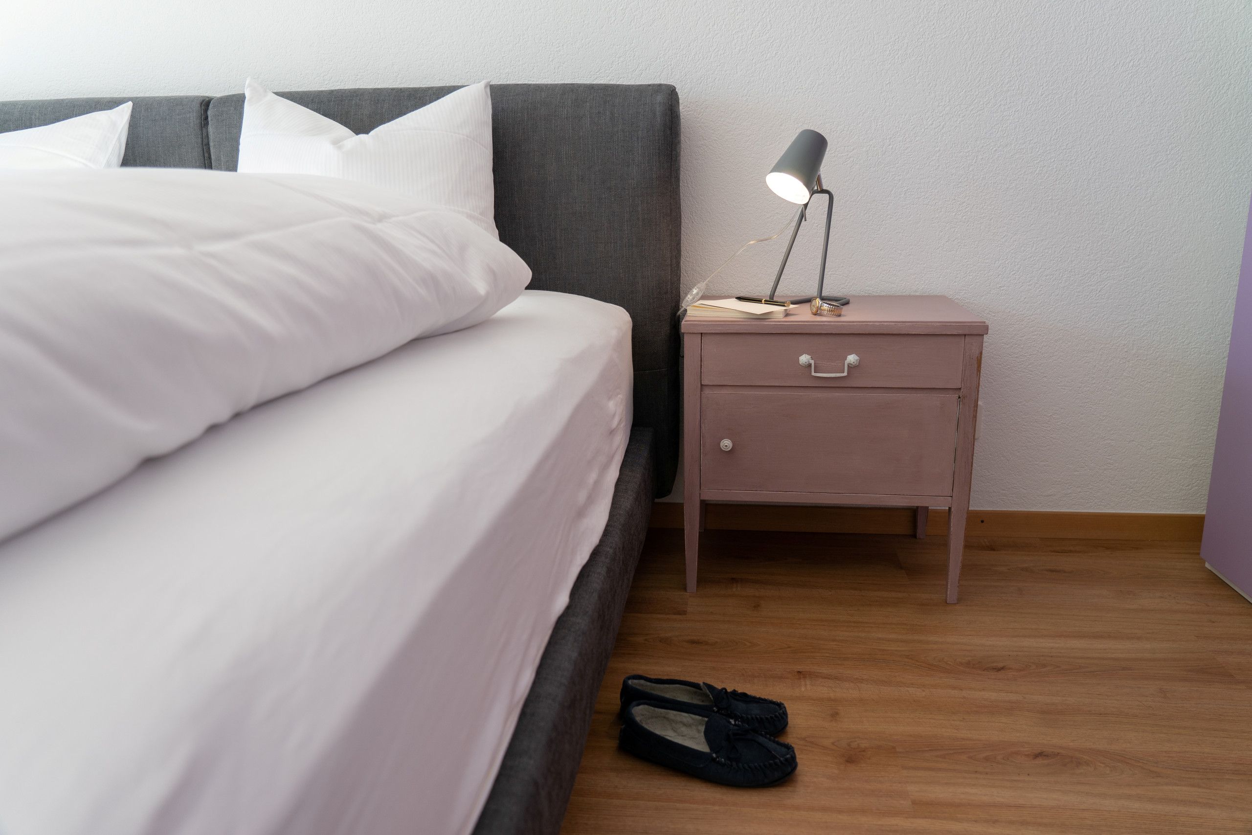 Un lit bien fait, avec du linge de lit blanc, une petite table de chevet avec une lampe, et une paire de chaussures posées sur le sol.