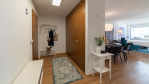 A modern hotel room with a wooden wardrobe, a small white table with a plant, a dining area with a round table and chairs, and a large window providing a view of the outdoors.