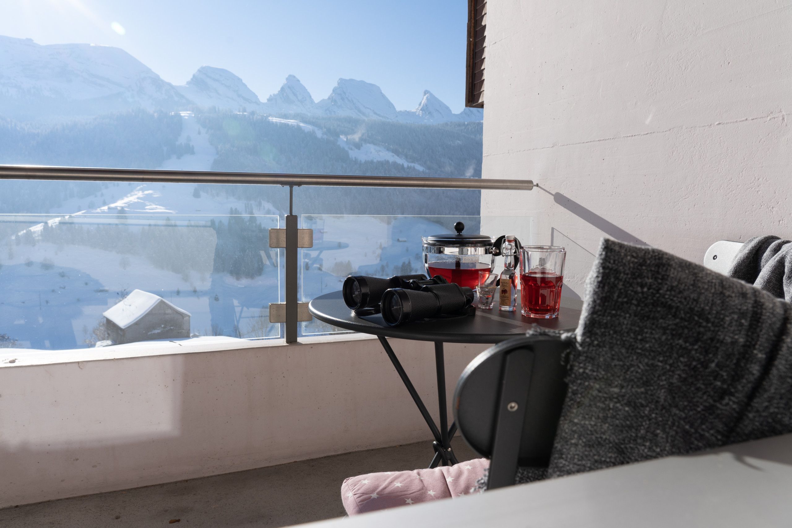 A balcony with a mountain view, a table with a camera, a glass of red liquid, and a bottle.