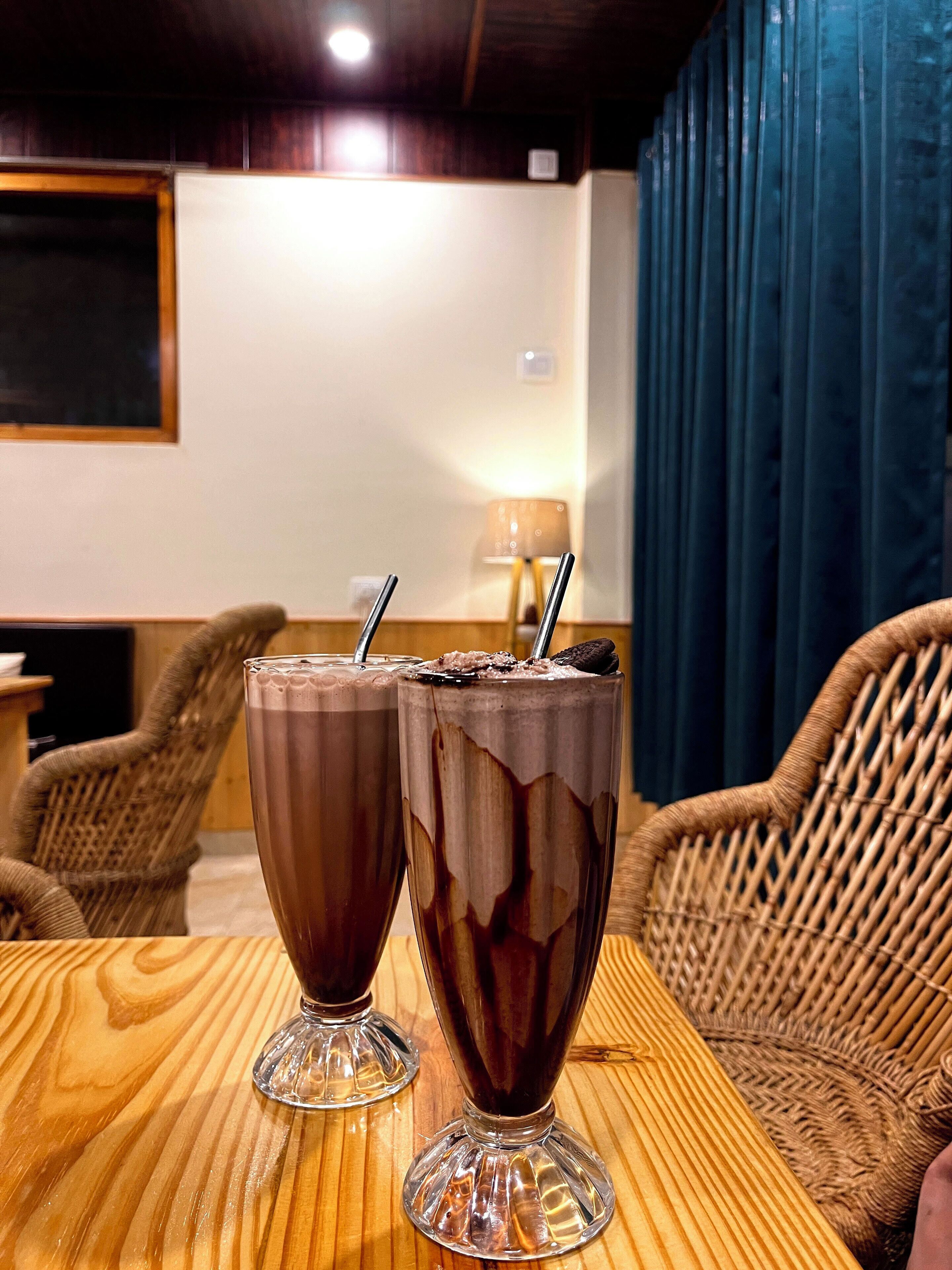 Deux milk-shakes au chocolat sur une table en bois, dans une pièce meublée de meubles en osier et ornée d’un rideau bleu.