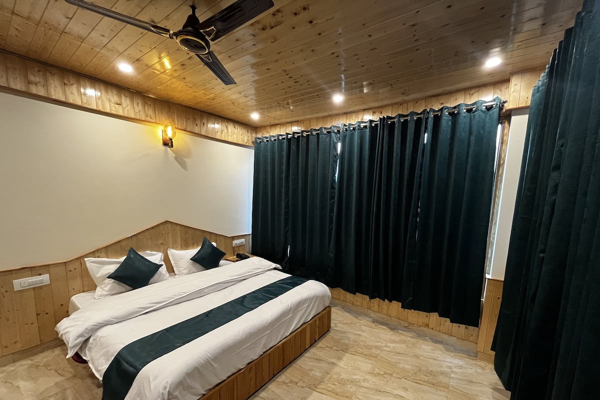 Un dormitorio con cabecera de madera, cama con ropa de cama blanca y verde, ventilador de techo y cortinas oscuras.