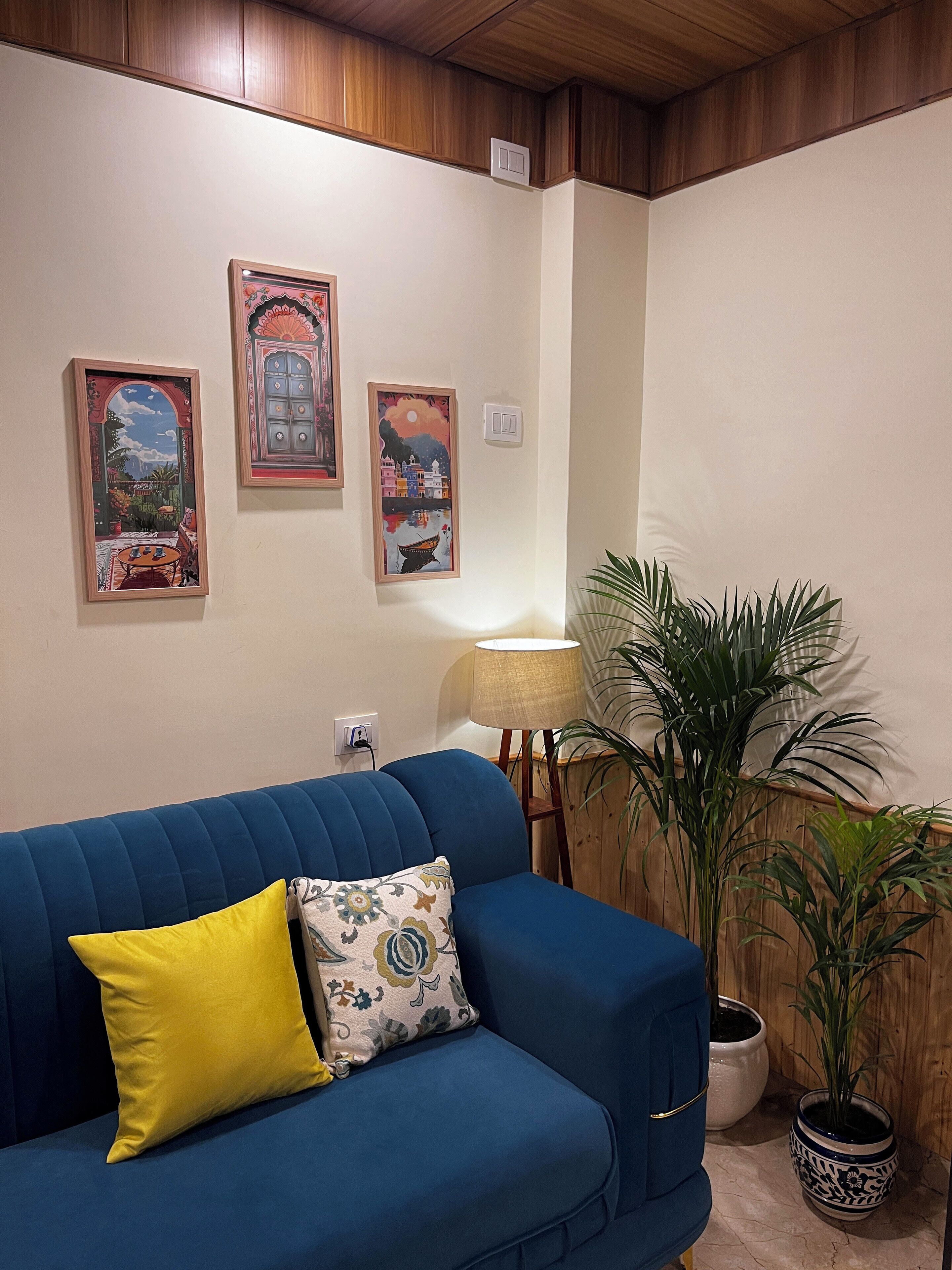A blue sofa with a yellow pillow, a lamp, and two framed artworks on the wall.