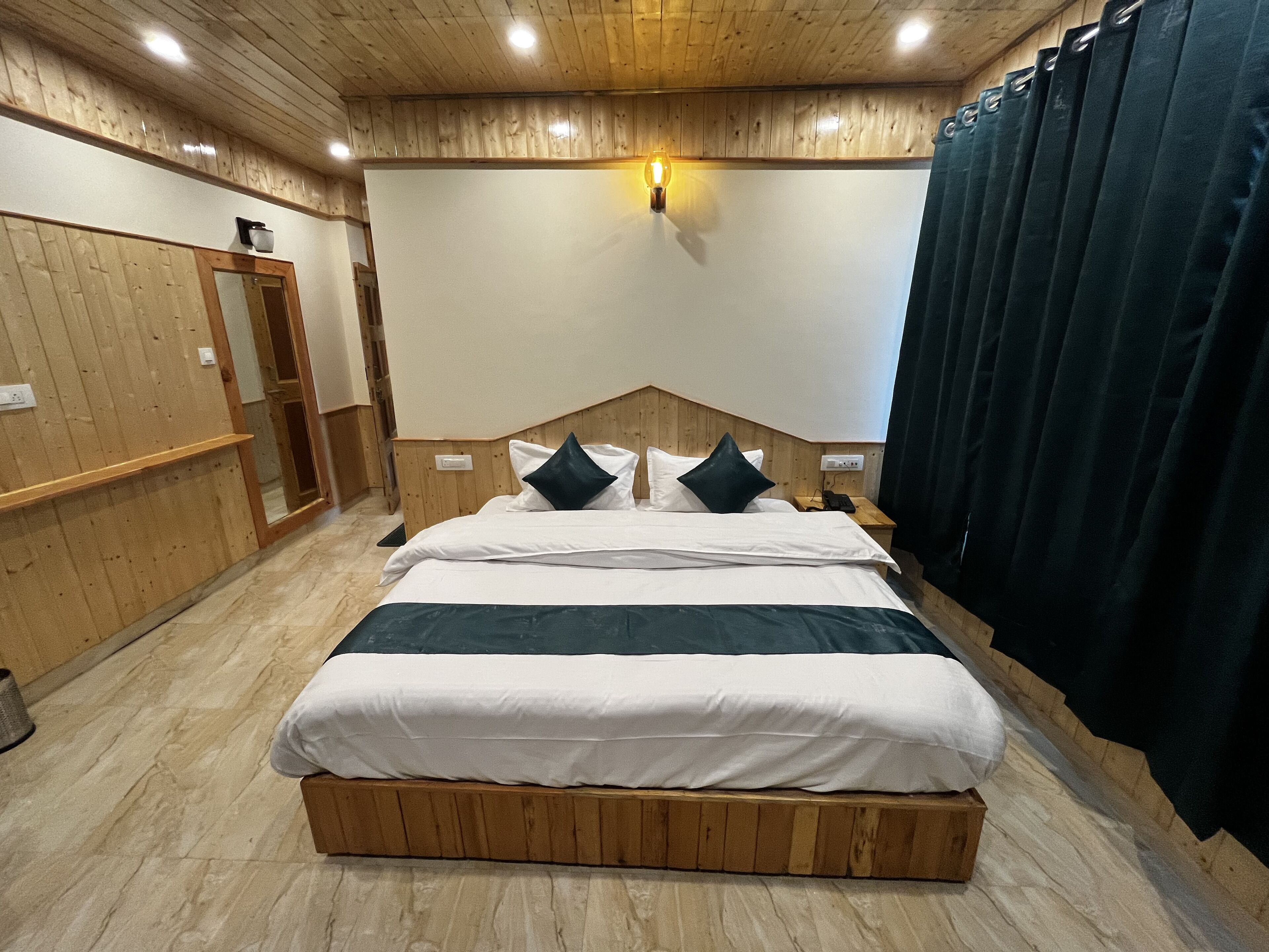 A bedroom with a wooden headboard, a large bed with white and dark green bedding, a bedside table with a lamp, and a wooden floor.