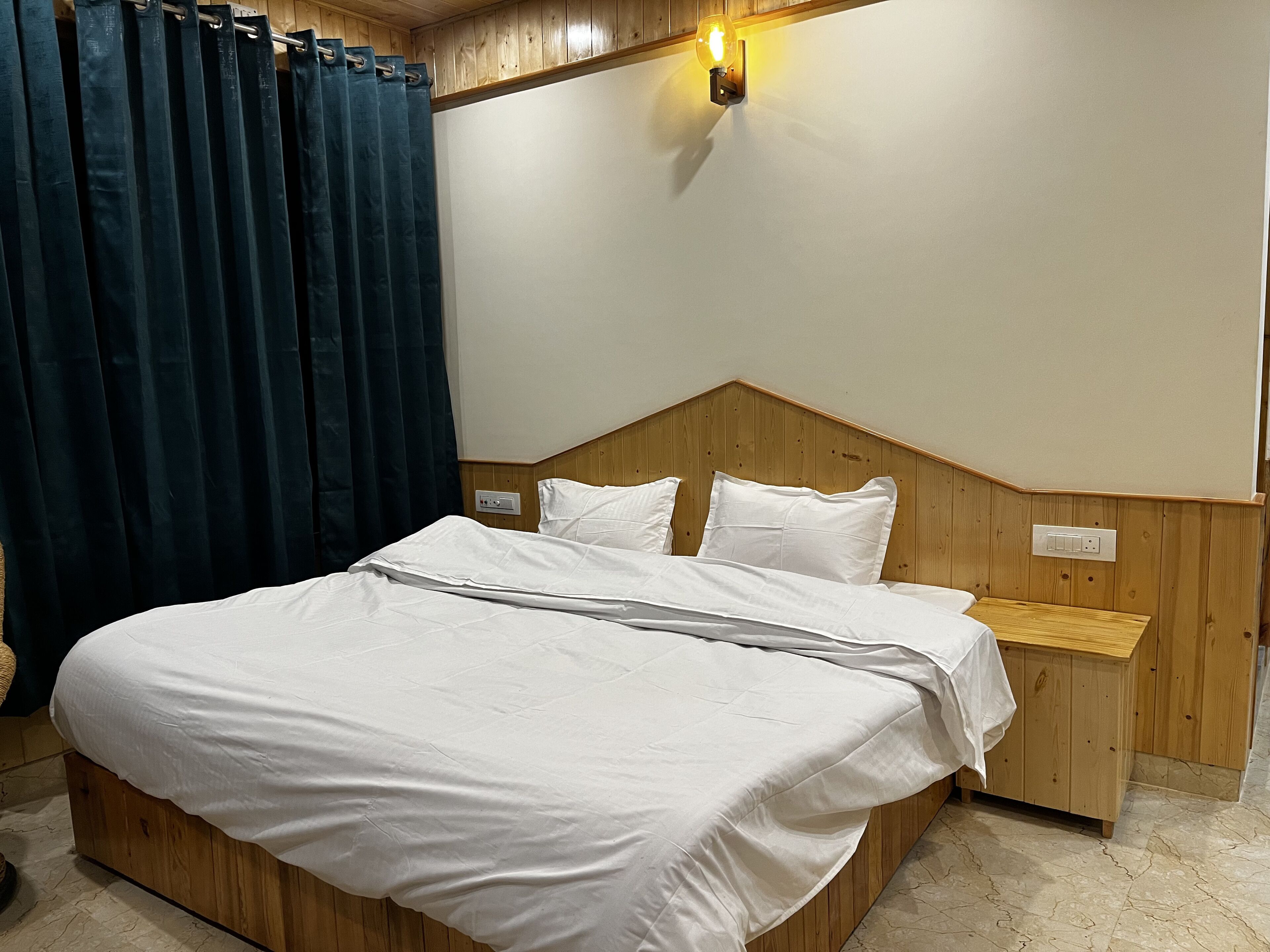 A bedroom with a wooden headboard, a bed with white bedding, a nightstand, a wall-mounted light, and a dark blue curtain.