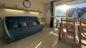 A cozy room with a blue sofa, a wooden dining table with chairs, a wall clock, and a window with a view of a snowy landscape. - Studio for 4, near chairlift, WiFi, parking - SuperDévoluy (Le Dévoluy)