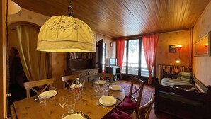 A cozy dining area with a wooden table set for four, a hanging lamp, and a view of the outdoors through large windows.