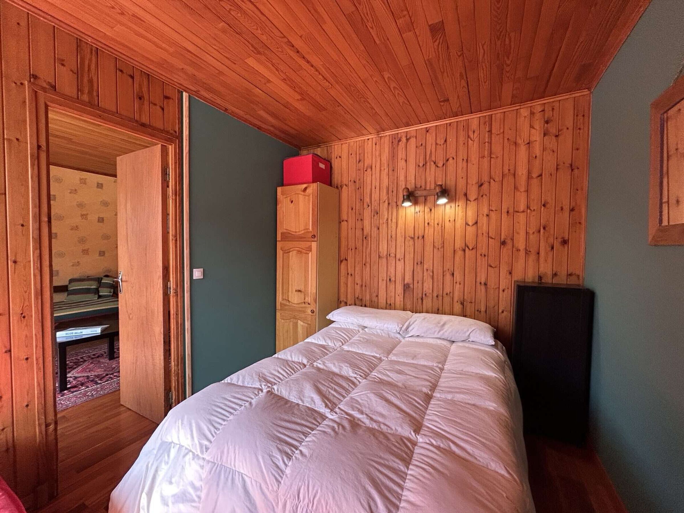A bedroom with wooden paneling, a bed with a white duvet, a red box on top of a wardrobe, and a speaker on the right.