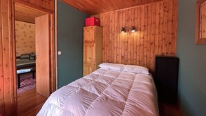 A bedroom with wooden paneling, a bed with a white duvet, a red box on top of a wardrobe, and a speaker on the right.