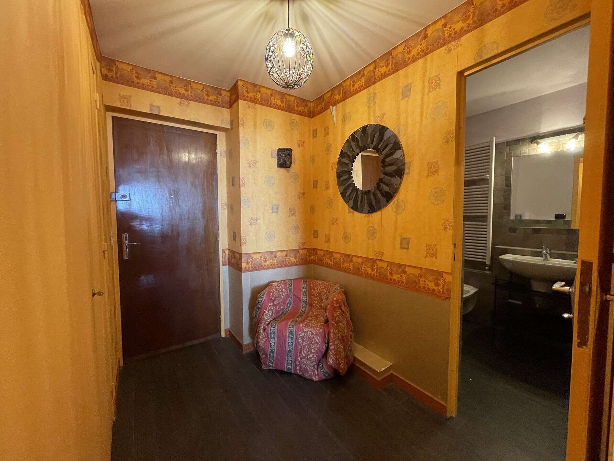 Un pasillo con puerta de madera, pared con diseño, espejo decorativo y un baño con lavamanos y espejo.