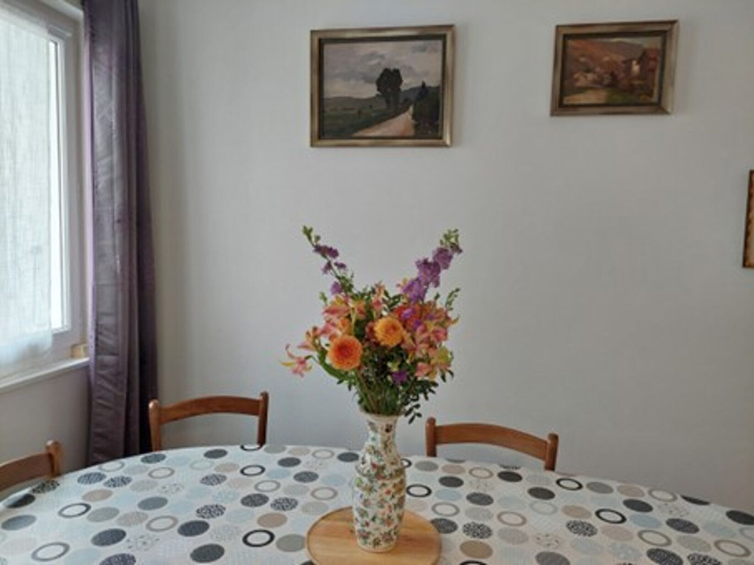 A dining table with a floral centerpiece, surrounded by chairs, in a room with two framed pictures on the wall.