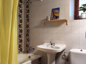 Une salle de bain avec une baignoire, un lavabo et des toilettes. On y trouve un rideau de douche à motifs floraux, une étagère avec un livre et une plante en pot.